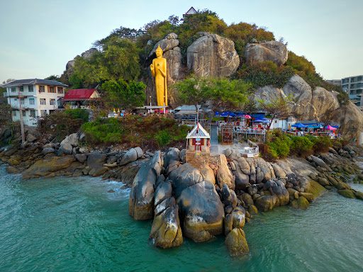 unnamed-3 a coastal view in Hua Hin with a towering golden Buddha statue standing on a rocky outcrop overlooking the sea