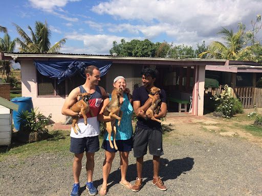 unnamed-9 Three smiling volunteers standing outside a house, each holding a puppy